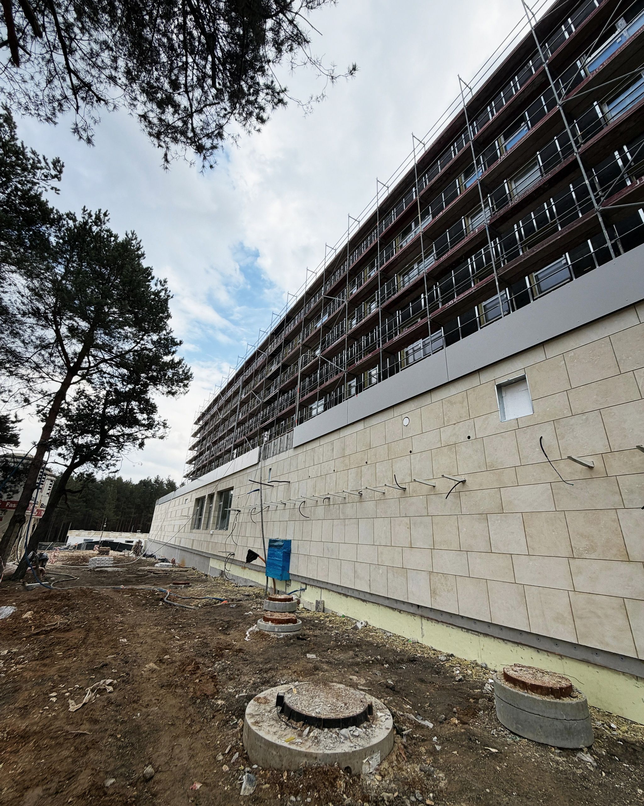 Elewacja z jasnych płyt kamiennych, wyższe kondygnacje w rusztowaniach, obok sosny