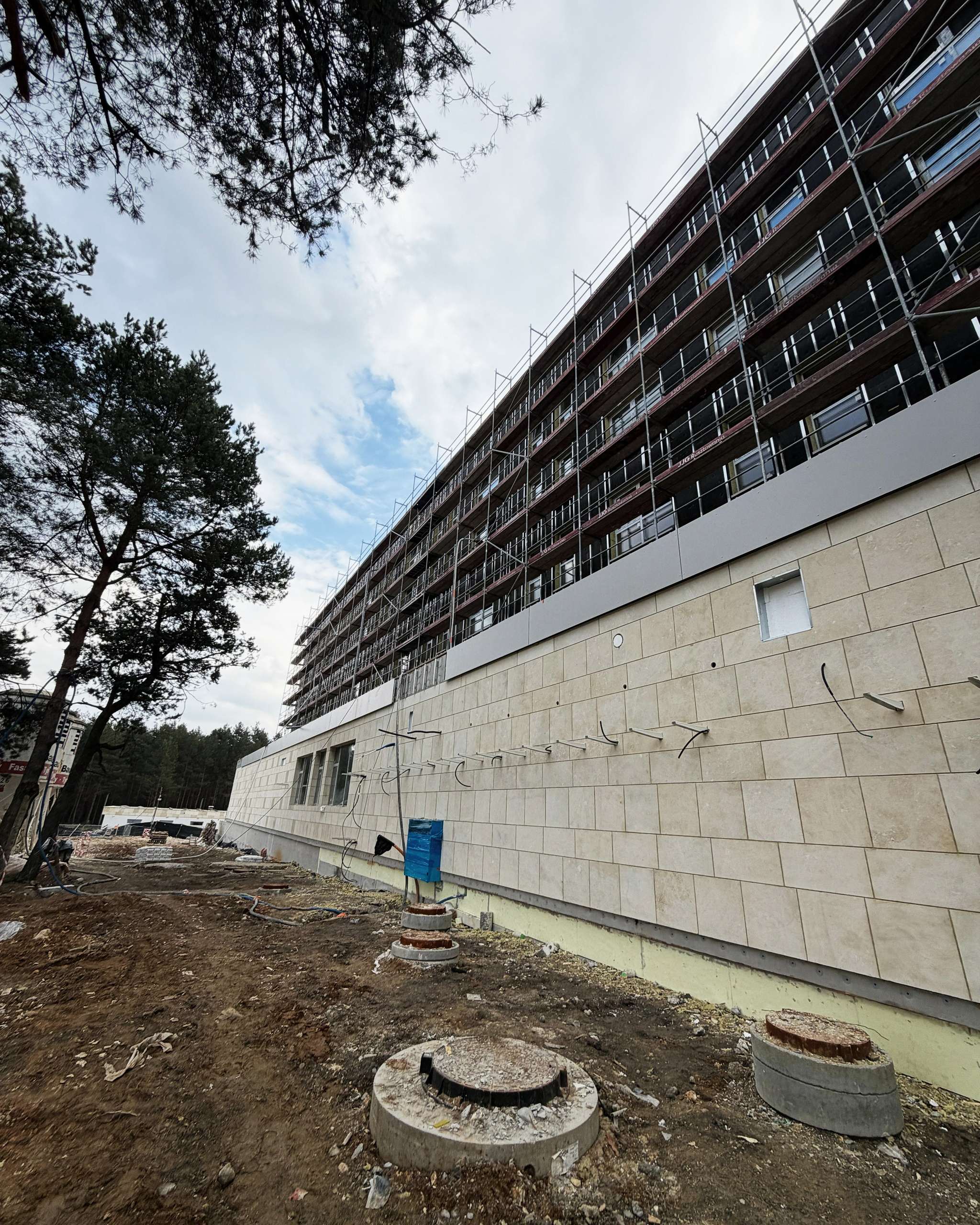 Elewacja z jasnych płyt kamiennych, wyższe kondygnacje w rusztowaniach, obok sosny